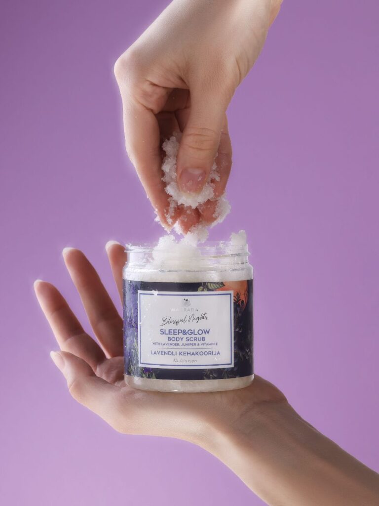 Lavender body scrub by Magrada Organic Cosmetics held in hand with scrub being scooped against purple background