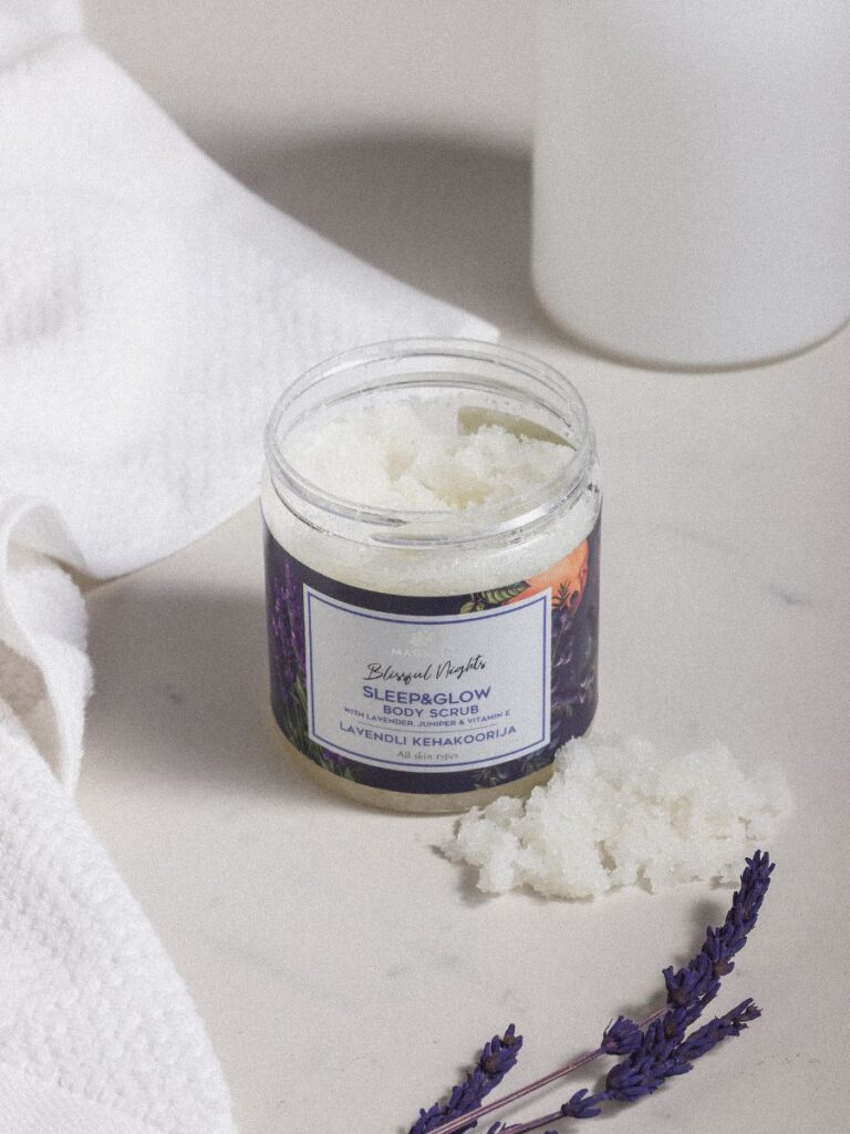 Magrada Organic Cosmetics lavender body scrub in a jar with scrub spilled beside it and lavender sprigs on white surface